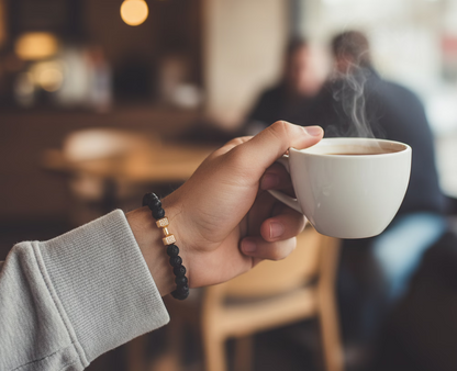 Hand wearing a natural stone bracelet holding a steaming white mug in a blurred café setting
